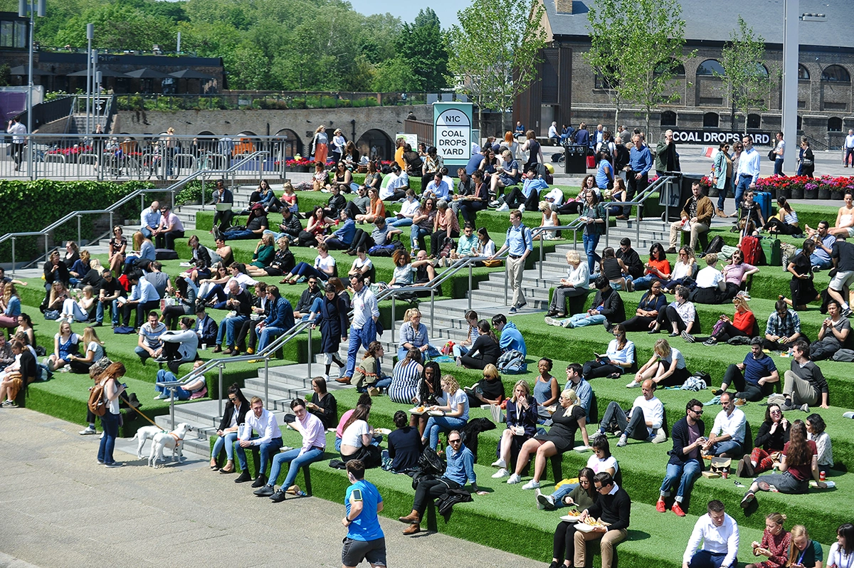 A large crowd of people sitting and standing on outdoor seating in a city public space on a sunny day.