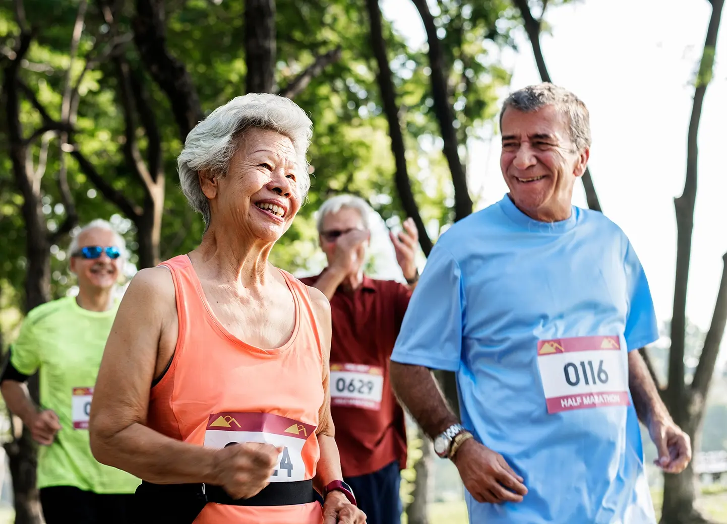 Two adults smiling while jogging in a running race, with other runners and trees in the background.